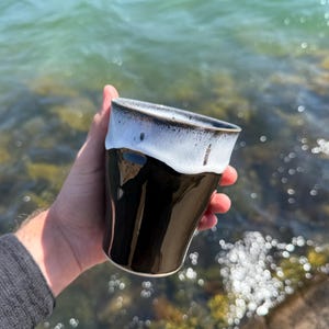 Könnte beinhalten: Eine schwarze Keramiktasse mit weißem und dunkelbraunem Rand. Die Tasse hat eine konische Form und wird von einer Hand gehalten. Der Hintergrund zeigt ein Gewässer und Felsen.