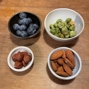 May include: Four small ceramic bowls containing food items. The top left bowl is black and holds fresh blueberries. The top right bowl is white and contains green wasabi peas. The bottom left bowl is white with hazelnuts, and the bottom right bowl is white with almonds.