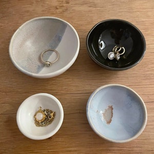 May include: Four small, round ceramic bowls of varying sizes and colors. One bowl is white with a grey wash, one is solid black, one is plain white, and one is light blue with a textured center. Each bowl contains a piece of jewelry.