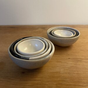May include: Two sets of nested ceramic bowls, each containing three bowls of varying sizes. The bowls feature a smooth, glossy interior in off-white, while the exterior surfaces display a mix of matte grey, black, and speckled grey finishes.