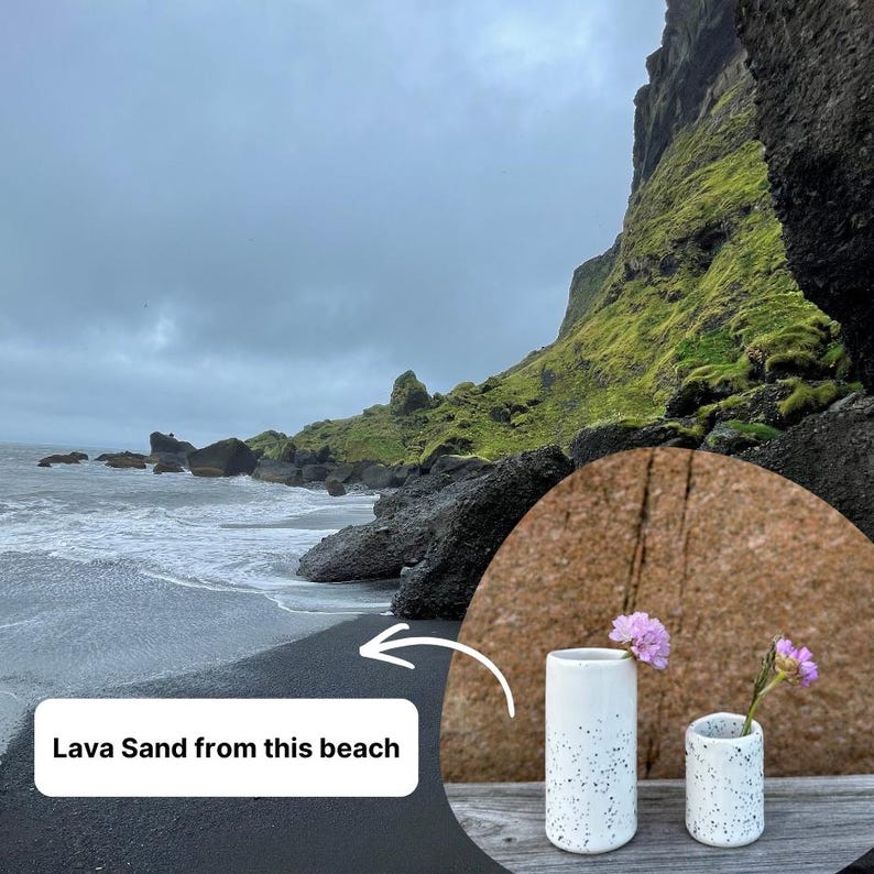 May include: Two cylindrical white ceramic vases with black speckles, each containing a single small pink flower. The image includes a text overlay stating Lava Sand from this beach, pointing to a coastal scene with black sand and green cliffs.