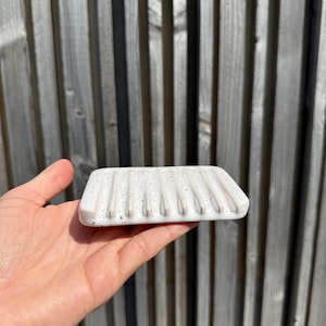 May include: A rectangular white soap dish featuring a speckled pattern with small black dots. The surface of the dish is designed with a series of parallel raised ridges that run across the width, creating channels for drainage and airflow.