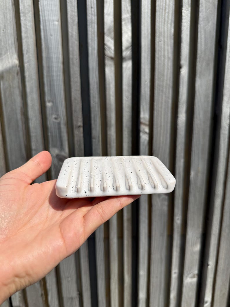 May include: A rectangular white soap dish featuring a speckled pattern with small black dots. The surface of the dish is designed with a series of parallel raised ridges that run across the width, creating channels for drainage and airflow.