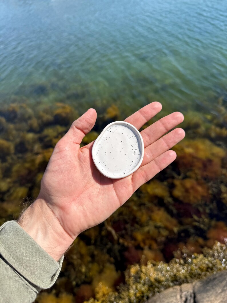 May include: A small, white, organic-shaped ceramic dish featuring a speckled pattern with tiny black dots. The dish has a smooth, slightly raised rim and a shallow, concave center, designed to hold small items like jewelry or trinkets.