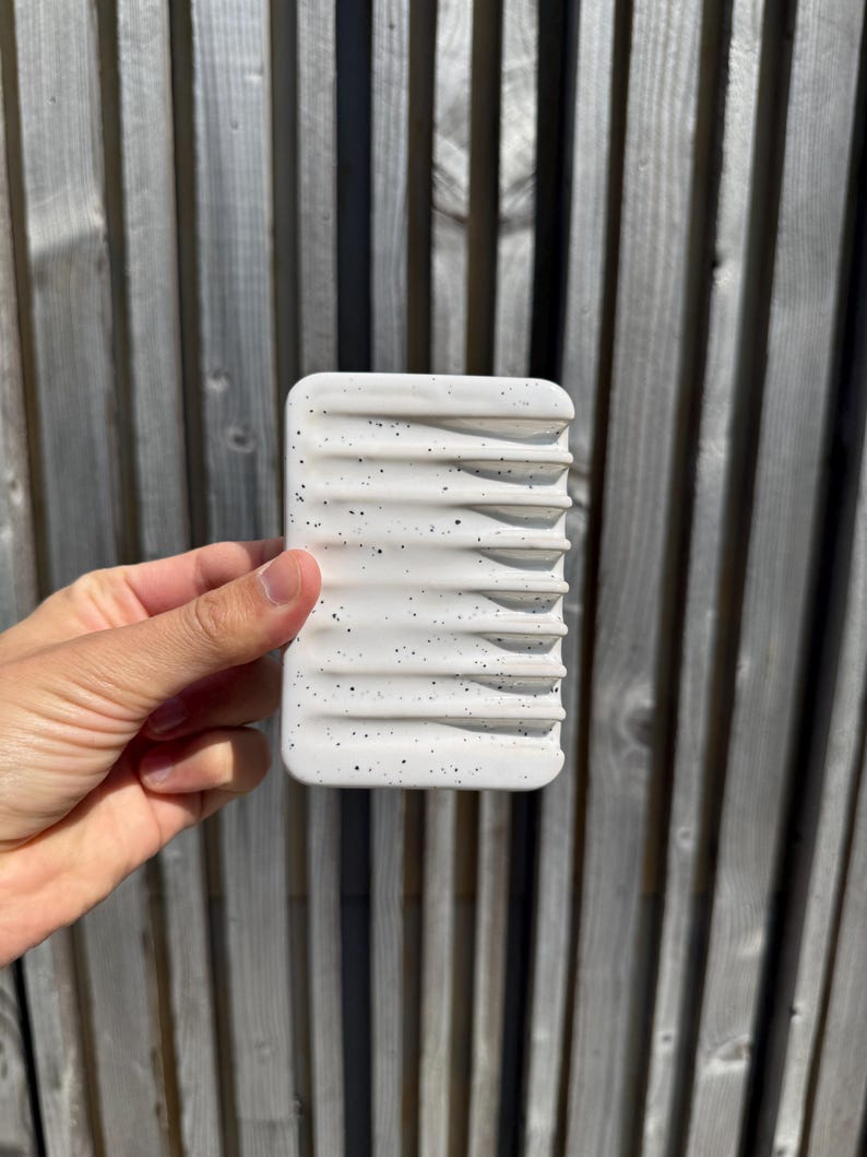 May include: A rectangular white soap dish featuring a speckled pattern of small black dots across its surface. The dish has a series of parallel horizontal ridges designed to elevate an object, with rounded corners and a smooth, matte finish.