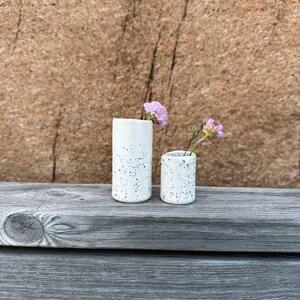 May include: Two cylindrical ceramic vases with a white glaze and black speckled pattern. The taller vase holds a single light purple flower, while the shorter vase contains a small purple flower and a thin green stem, creating a simple display.