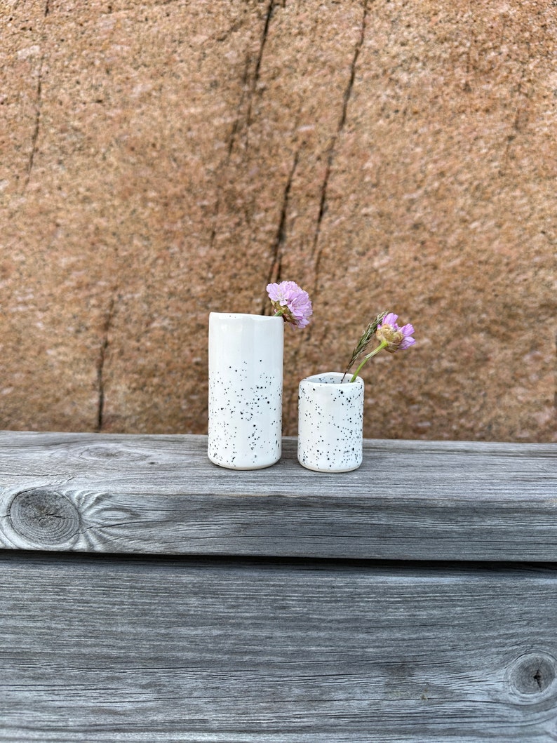 May include: Two cylindrical ceramic vases with a white glaze and black speckled pattern. The taller vase holds a single light purple flower, while the shorter vase contains a small purple flower and a thin green stem, creating a simple display.