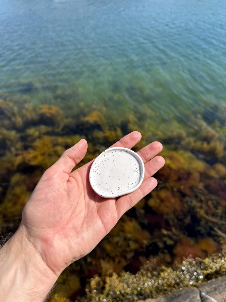 May include: A small, round, white ceramic trinket dish with an irregular, organic shape and a speckled pattern featuring tiny black dots scattered across the smooth surface. The dish has a slightly raised rim and a glossy finish.