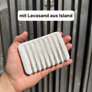 May include: A rectangular white soap dish featuring vertical drainage grooves and black speckles, with the text "mit Lavasand aus Island" printed above it. The dish has a smooth finish and a series of parallel channels for water drainage.