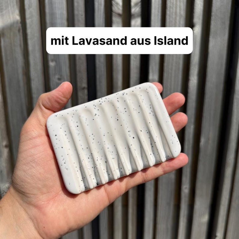 May include: A rectangular white soap dish featuring vertical drainage grooves and black speckles, with the text "mit Lavasand aus Island" printed above it. The dish has a smooth finish and a series of parallel channels for water drainage.