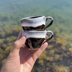 Könnte beinhalten: Zwei kleine Espresso-Tassen aus Keramik, gestapelt, in einer Hand gehalten. Die Tassen sind schwarz mit einer weißen und grauen Glasur am Rand. Jede Tasse hat einen kleinen Henkel. Der Hintergrund ist eine verschwommene Ansicht von Wasser und Vegetation.