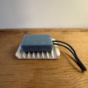 May include: A rectangular grey bar of soap with a dark grey rope loop attached, resting on a white ceramic soap dish featuring raised parallel ridges. The soap has a textured surface and the dish has a smooth, glossy finish.