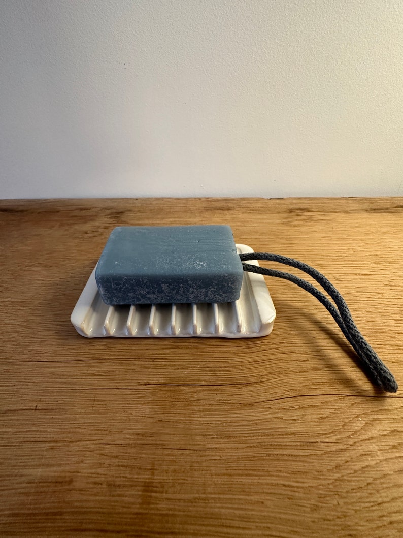 May include: A rectangular grey bar of soap with a dark grey rope loop attached, resting on a white ceramic soap dish featuring raised parallel ridges. The soap has a textured surface and the dish has a smooth, glossy finish.