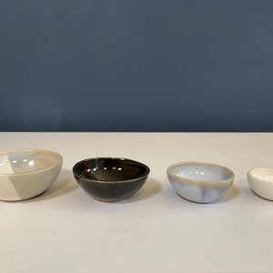 May include: A set of four small ceramic bowls arranged in a row. The first bowl is cream and light gray, the second is glossy black, the third is a mottled light blue and white, and the fourth is a small, solid cream-colored bowl with a smooth finish.