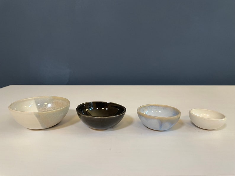 May include: A set of four small ceramic bowls arranged in a row. The first bowl is cream and light gray, the second is glossy black, the third is a mottled light blue and white, and the fourth is a small, solid cream-colored bowl with a smooth finish.