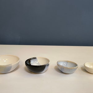 May include: A set of four ceramic bowls arranged in descending order of size. The bowls feature a mix of glossy white, light blue, and black glazes, with some showing a two-tone color application. Each bowl has a smooth, rounded rim and base.
