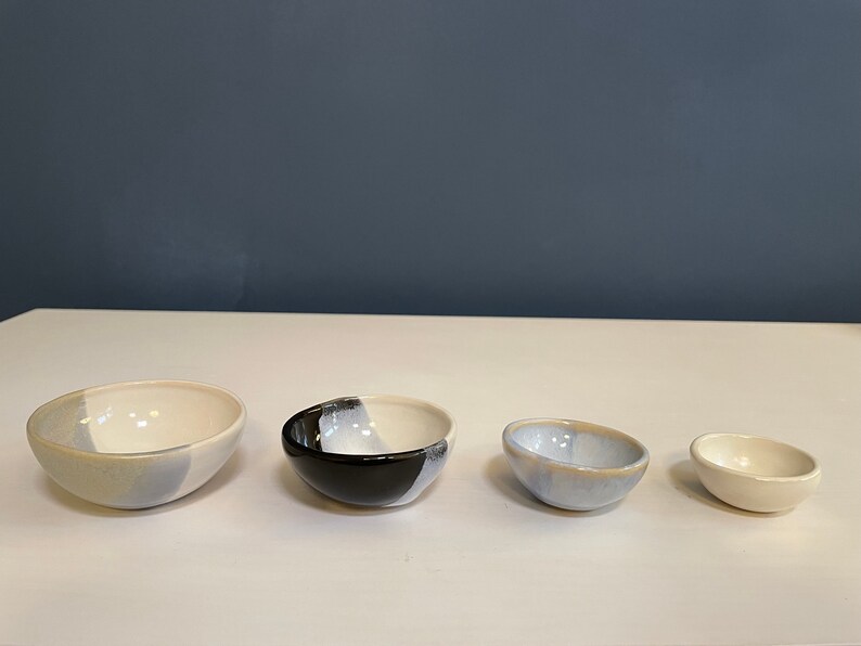 May include: A set of four ceramic bowls arranged in descending order of size. The bowls feature a mix of glossy white, light blue, and black glazes, with some showing a two-tone color application. Each bowl has a smooth, rounded rim and base.
