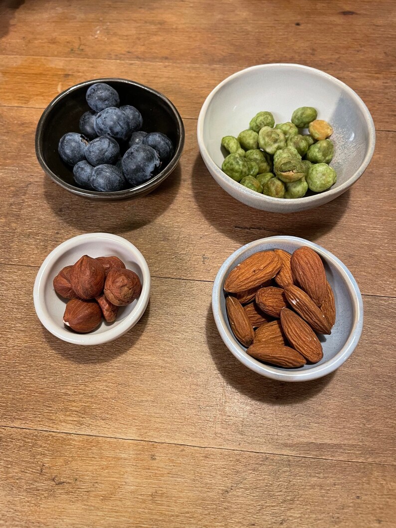 May include: Four small ceramic bowls containing food items. The top left bowl is black and holds fresh blueberries. The top right bowl is white and contains green wasabi peas. The bottom left bowl is white with hazelnuts, and the bottom right bowl is white with almonds.