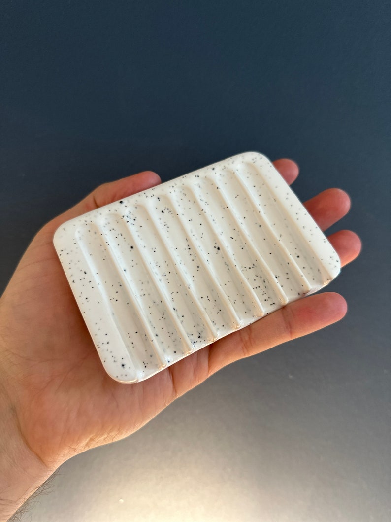 May include: A rectangular white ceramic soap dish featuring a speckled pattern with small black dots. The surface of the dish is designed with a series of parallel, raised ridges that run across the width, creating channels for drainage.