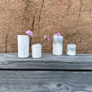 May include: Four ceramic vessels of varying heights and widths. Two are white with black speckles, and two feature a white base with a greyish-blue glazed top. Small purple flowers are placed inside two of the vessels, which are cylindrical in shape.