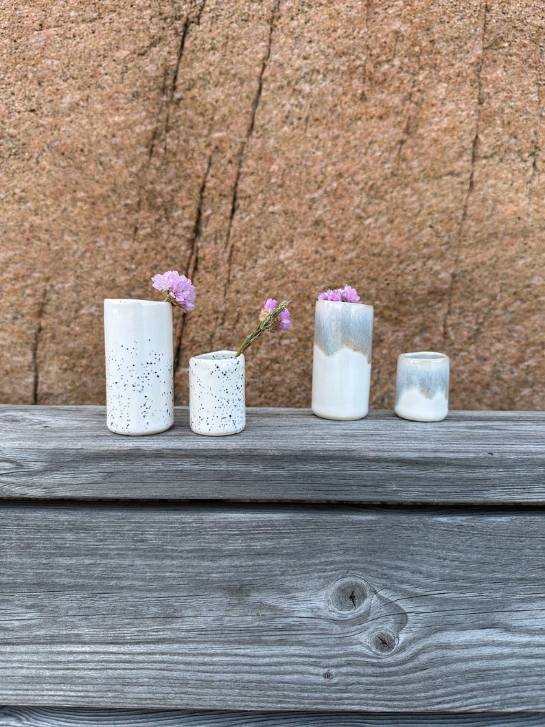 May include: Four ceramic vessels of varying heights and widths. Two are white with black speckles, and two feature a white base with a greyish-blue glazed top. Small purple flowers are placed inside two of the vessels, which are cylindrical in shape.