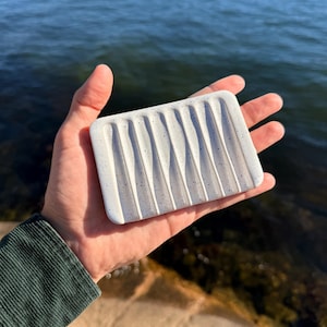 May include: A rectangular white soap dish featuring a speckled pattern with small blue dots and a series of vertical, parallel grooves carved into the surface to allow for drainage, held in a palm against a blurred water background.