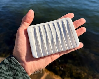 Op de afbeelding: Een rechthoekig wit zeepbakje met een gespikkeld patroon van kleine blauwe stippen en een reeks verticale, parallelle groeven in het oppervlak die zijn aangebracht om drainage mogelijk te maken, vastgehouden in een handpalm.
