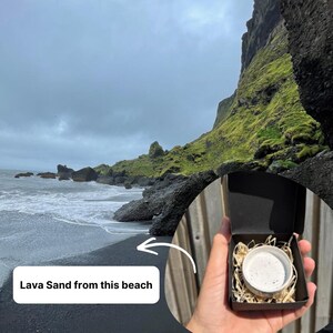 May include: A small black cardboard box containing a white ceramic container filled with dark speckled lava sand, nestled in light-colored wood shavings. The box is held against a backdrop of a black sand beach with mossy cliffs and a cloudy sky.