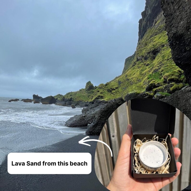 May include: A small black cardboard box containing a white ceramic container filled with dark speckled lava sand, nestled in light-colored wood shavings. The box is held against a backdrop of a black sand beach with mossy cliffs and a cloudy sky.