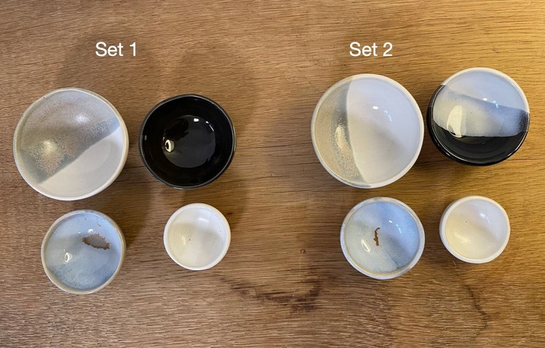 May include: Two sets of ceramic bowls labeled Set 1 and Set 2. Each set contains four bowls of varying sizes and finishes, including solid black, white, and blue-grey glazed interiors. The bowls are arranged in two rows of two, showing circular rims.