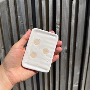 May include: A rectangular white ceramic soap dish with a glossy finish, featuring a speckled pattern of small black dots and three circular beige indentations on the surface. The dish has a slightly wavy, ridged texture across its top surface.