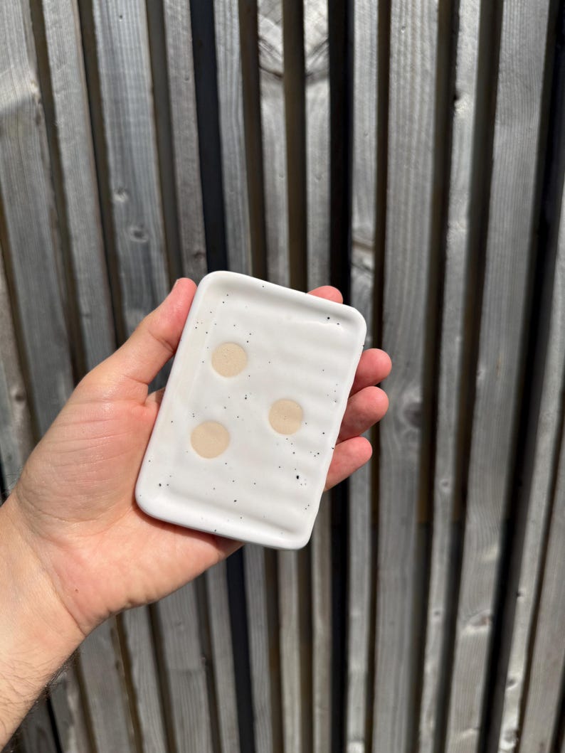 May include: A rectangular white ceramic soap dish with a glossy finish, featuring a speckled pattern of small black dots and three circular beige indentations on the surface. The dish has a slightly wavy, ridged texture across its top surface.