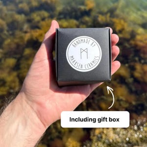 May include: A square black gift box featuring a circular white sticker on the front. The sticker displays the text "HANDMADE BY MAAKSEN CERAMICS" in a circular arrangement around a central geometric rune-like symbol. The box is held in a palm.
