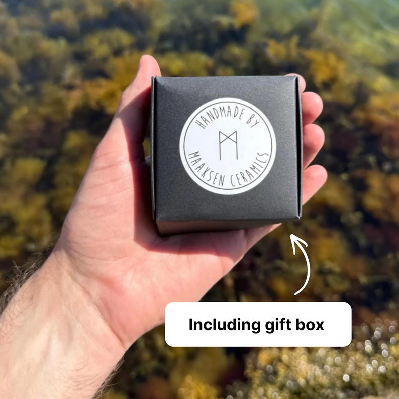 May include: A square black gift box featuring a circular white sticker on the front. The sticker displays the text "HANDMADE BY MAAKSEN CERAMICS" in a circular arrangement around a central geometric rune-like symbol. The box is held in a palm.