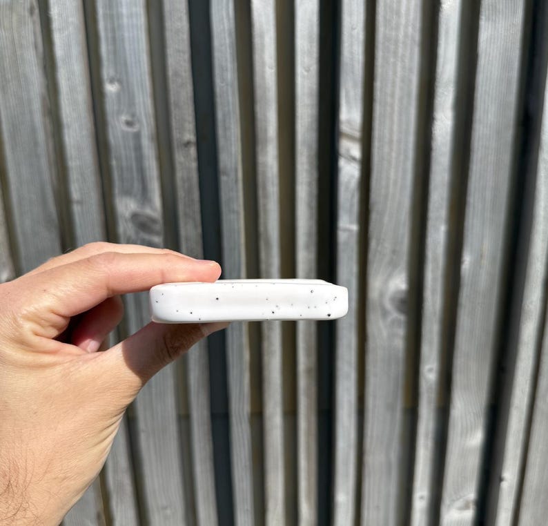 May include: A rectangular white ceramic object with a speckled pattern featuring small black dots across its surface. The object has rounded edges and a smooth, matte finish, presented in a horizontal orientation with a visible thickness.