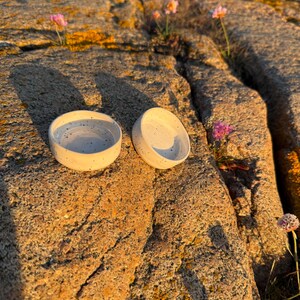 May include: Two small, round, off-white ceramic bowls featuring a speckled pattern with dark grey dots. The bowls have a smooth, glazed finish and a simple, low-profile design. They are positioned side-by-side on a textured, uneven rock surface.
