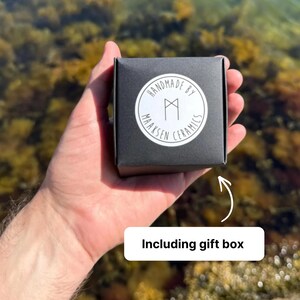 K&ouml;nnte beinhalten: Eine quadratische schwarze Geschenkbox mit einem runden wei&szlig;en Aufkleber auf der Vorderseite. Der Aufkleber zeigt den Text "HANDMADE BY MAAKSEN CERAMICS" in kreisf&ouml;rmiger Anordnung um ein zentrales geometrisches Symbol. Die Box liegt in einer Handfl&auml;che.