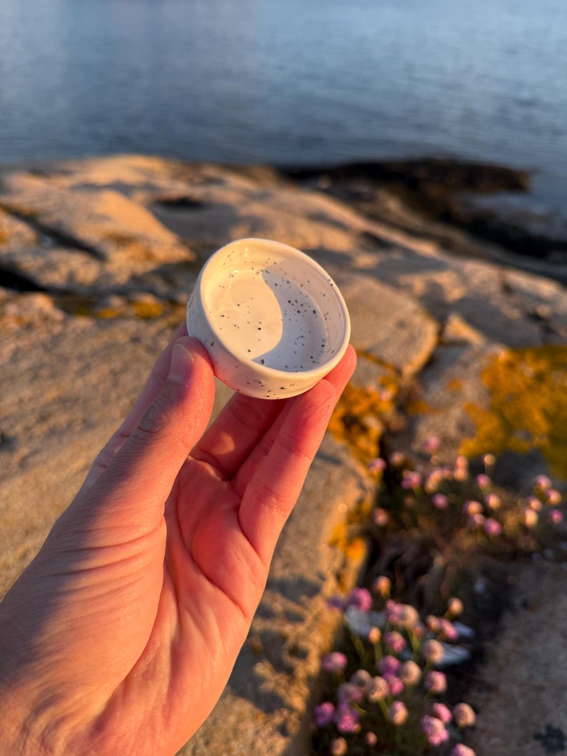 May include: A small, round, white ceramic dish featuring a speckled pattern with tiny black dots across its surface. The dish has a smooth, slightly curved interior and a raised rim, presenting a simple, functional design for holding small items.