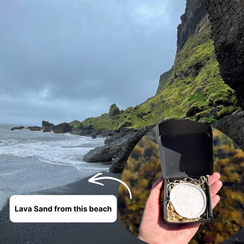 May include: A small black cardboard box containing a white, speckled, circular ceramic dish nestled in light-colored wood shavings. The box is held against a background of a black sand beach with mossy cliffs and the text Lava Sand from this beach.