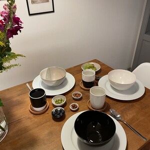 May include: A dining table setting featuring three ceramic bowls in white and black, two matching two-tone mugs on cork coasters, and four small bowls containing blueberries, green grapes, almonds, and hazelnuts, accompanied by a silver spoon.