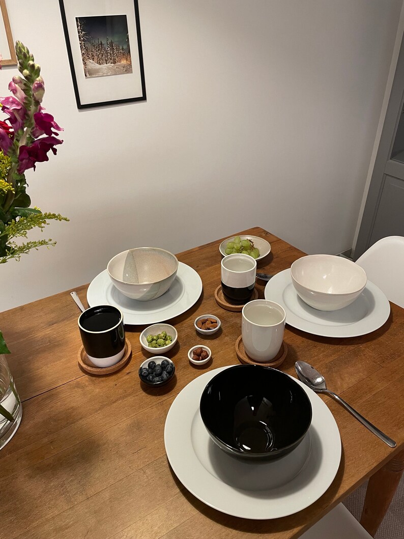 May include: A dining table setting featuring three ceramic bowls in white and black, two matching two-tone mugs on cork coasters, and four small bowls containing blueberries, green grapes, almonds, and hazelnuts, accompanied by a silver spoon.