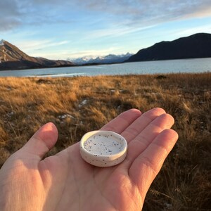 Op de afbeelding: Een kleine, ronde, gebroken witte keramische schaal met een gespikkeld patroon van kleine zwarte stippen over het oppervlak. De schaal heeft een ondiep, concaaf midden en een gladde, afgeronde rand, ontworpen als een compacte schaal.