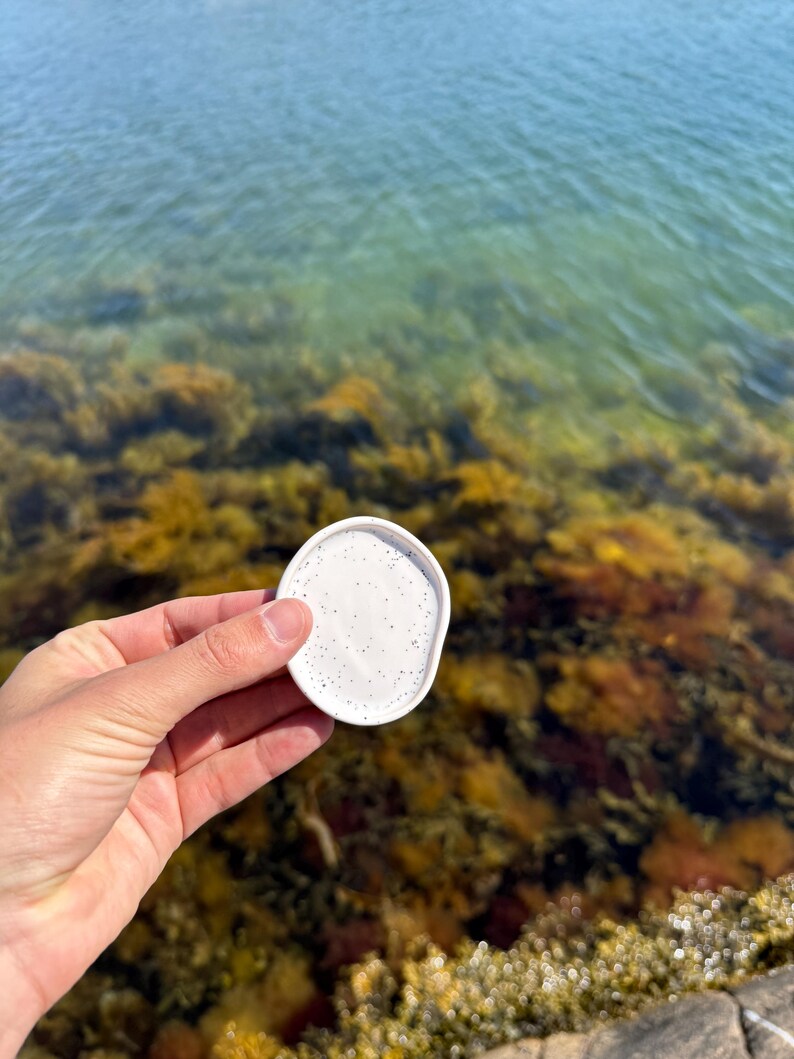 May include: A small, circular, white ceramic dish with a speckled pattern featuring tiny black dots. The dish has a slightly raised rim and a smooth, matte finish, presented as a simple, functional object for holding small items or trinkets.