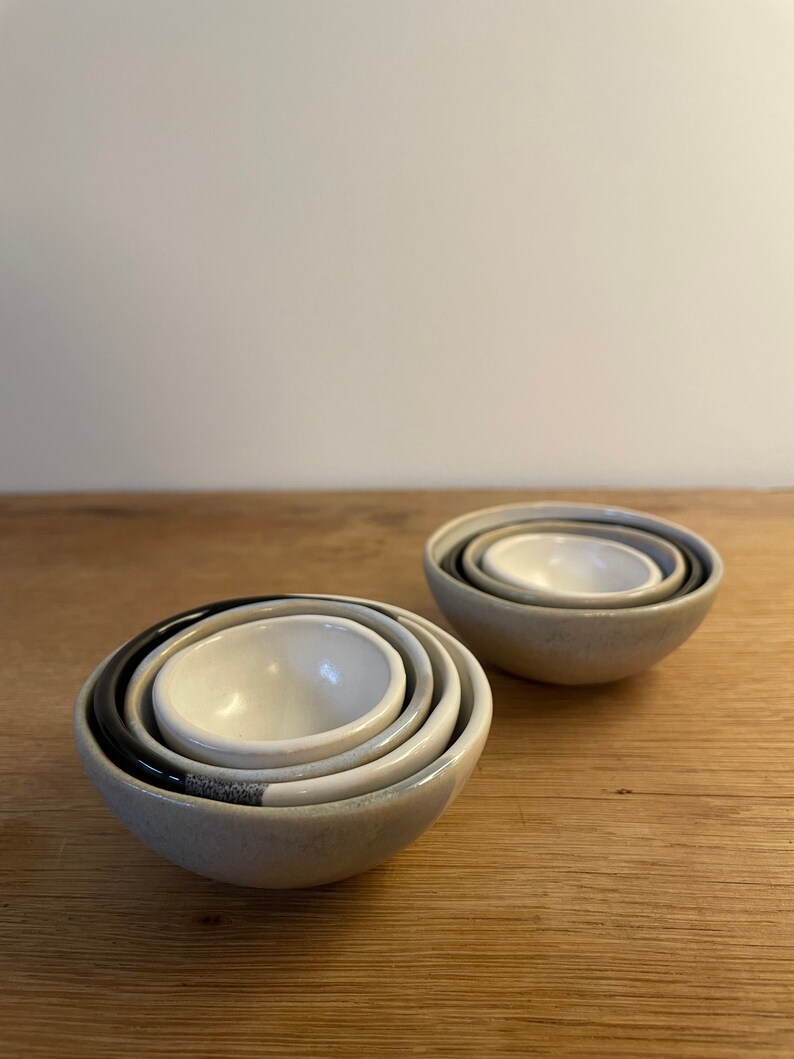 May include: Two sets of nested ceramic bowls, each containing three bowls of varying sizes. The bowls feature a smooth, glossy interior in off-white, while the exterior surfaces display a mix of matte grey, black, and speckled grey finishes.