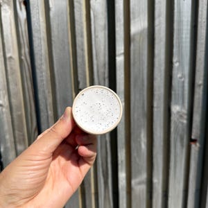 May include: A small, circular ceramic dish with a smooth, off-white glaze featuring a speckled pattern of tiny black dots across the surface. The dish has a simple, clean rim and a rounded edge, presenting a neutral and functional design.