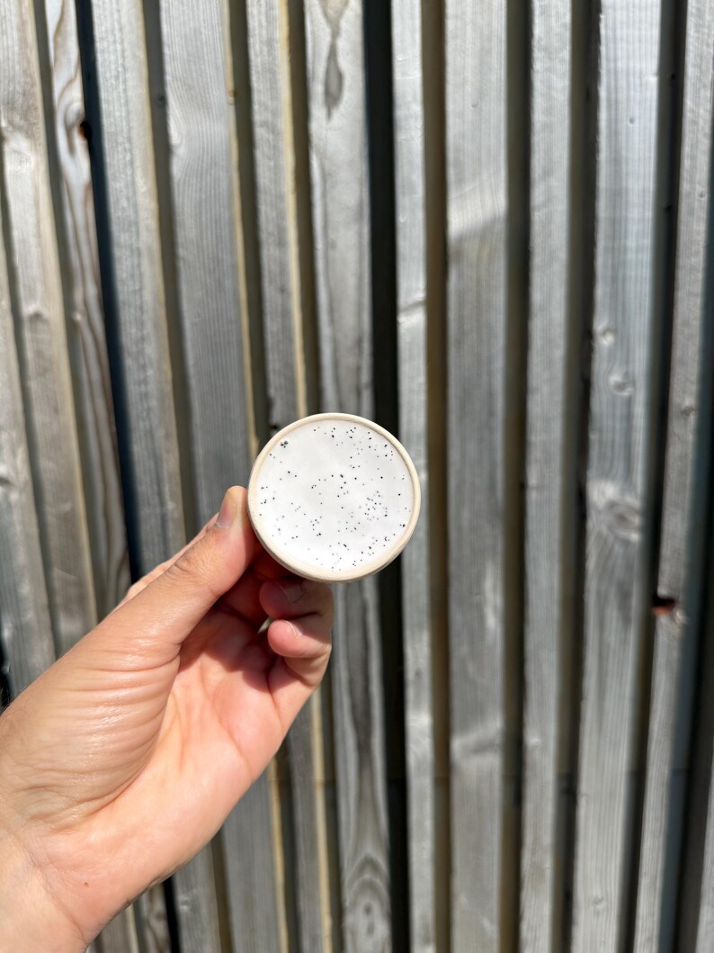 May include: A small, circular ceramic dish with a smooth, off-white glaze featuring a speckled pattern of tiny black dots across the surface. The dish has a simple, clean rim and a rounded edge, presenting a neutral and functional design.