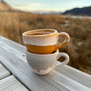 May include: Two ceramic coffee cups stacked on top of each other. The top cup is a light brown color with a white rim and a white stripe around the middle. The bottom cup is a light gray color with a white rim. The cups are sitting on a wooden surface.
