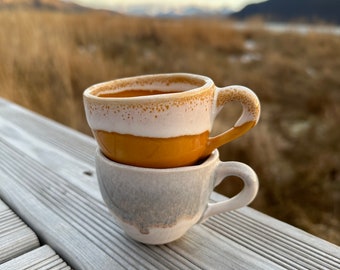 May include: Two ceramic coffee cups stacked on top of each other. The top cup is a light brown color with a white rim and a white stripe around the middle. The bottom cup is a light gray color with a white rim. The cups are sitting on a wooden surface.