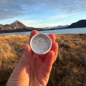 Op de afbeelding: Een klein, rond keramisch schaaltje met een witte glazuurlaag en een gespikkeld patroon van zwarte stippen. Het schaaltje heeft een gladde, afgeronde rand en een ondiepe binnenkant, wat zorgt voor een strak ontwerp met een contrasterende textuur.
