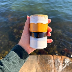 May include: A set of two stacked ceramic cups. The top cup features a white rim with a speckled texture transitioning into a solid mustard yellow base. The bottom cup has a glossy dark brown upper section and a smooth white lower base.